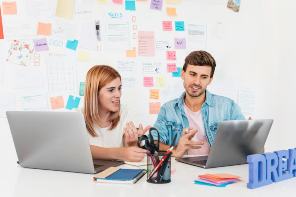 Two people sit at a white desk with laptops, engaged in discussion. A wall filled with colorful notes and papers is behind them, conveying creativity.