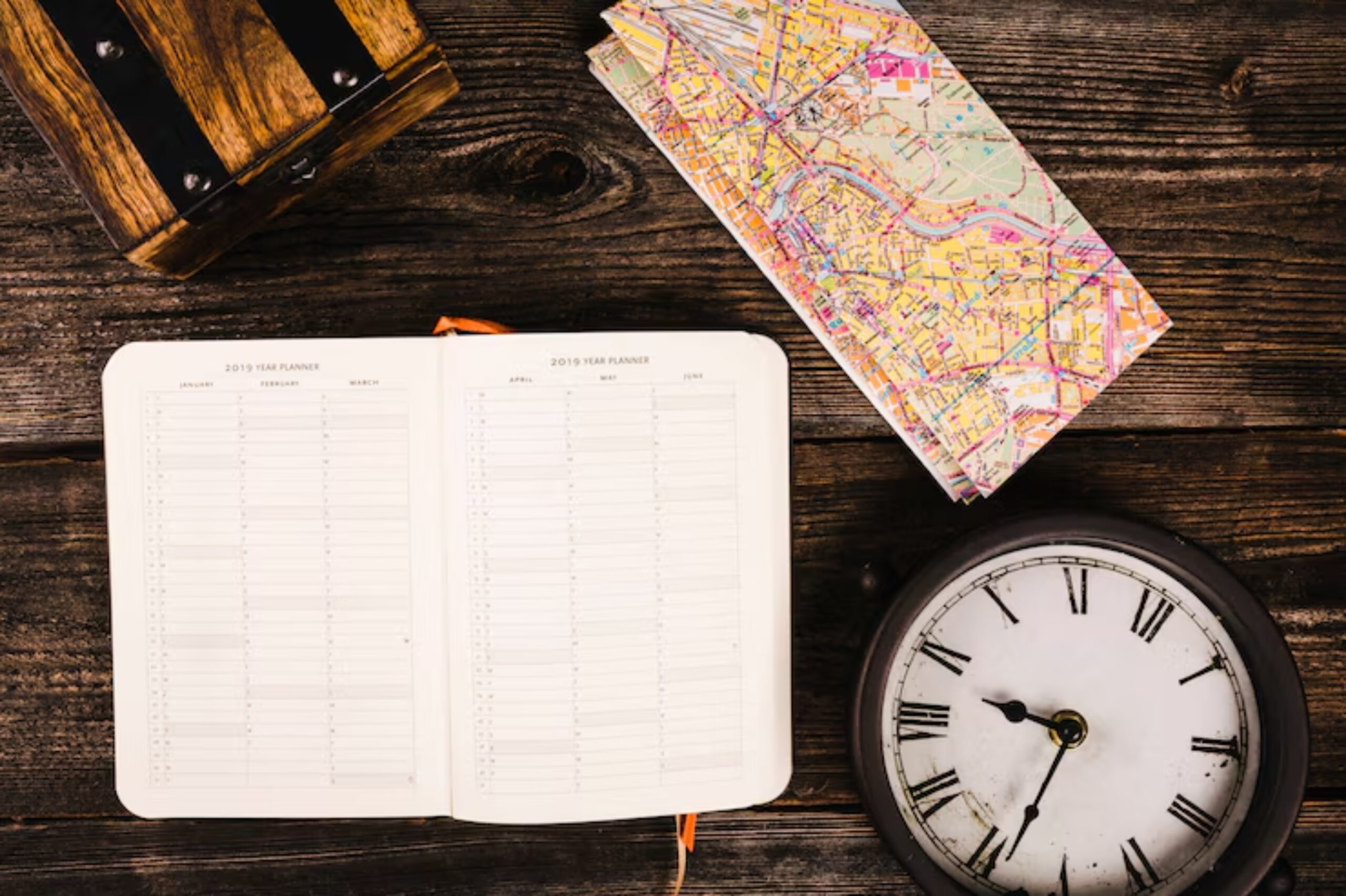 Open planner with calendar pages, a colorful map, and a vintage clock on rustic wooden table, evoking a sense of travel and planning.