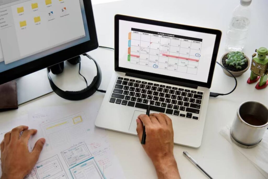 A person writes on paper while using a laptop to view a calendar, with a desktop monitor, a water bottle, and a plant nearby.