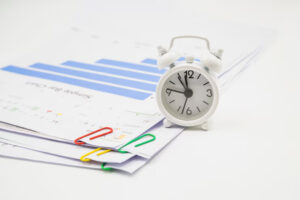 A white alarm clock rests on a stack of documents with colorful paperclips, featuring graphs and charts in a bright workspace.