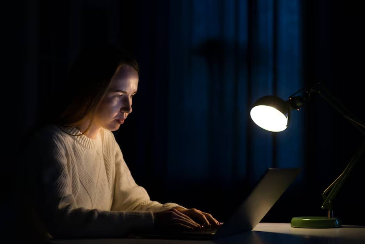 A person is typing on a laptop, illuminated by a desk lamp in a dimly lit room, creating a cozy night-time atmosphere.