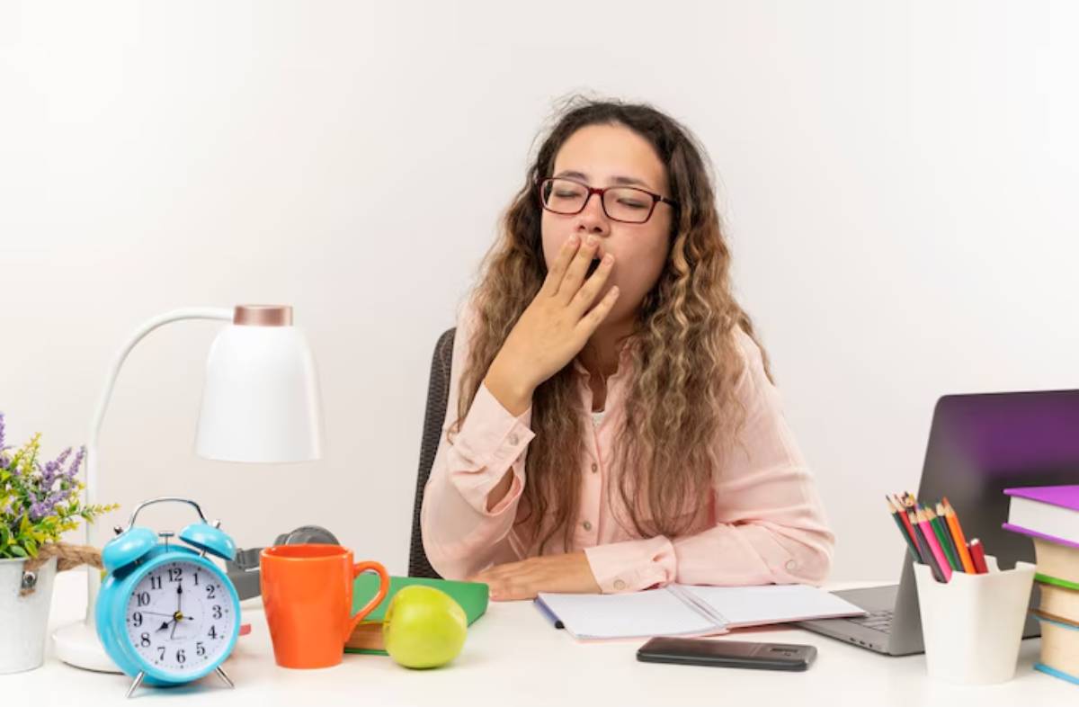 A woman with curly hair sits at a desk with a lamp, clock, books, snacks, and a laptop, appearing deep in thought.