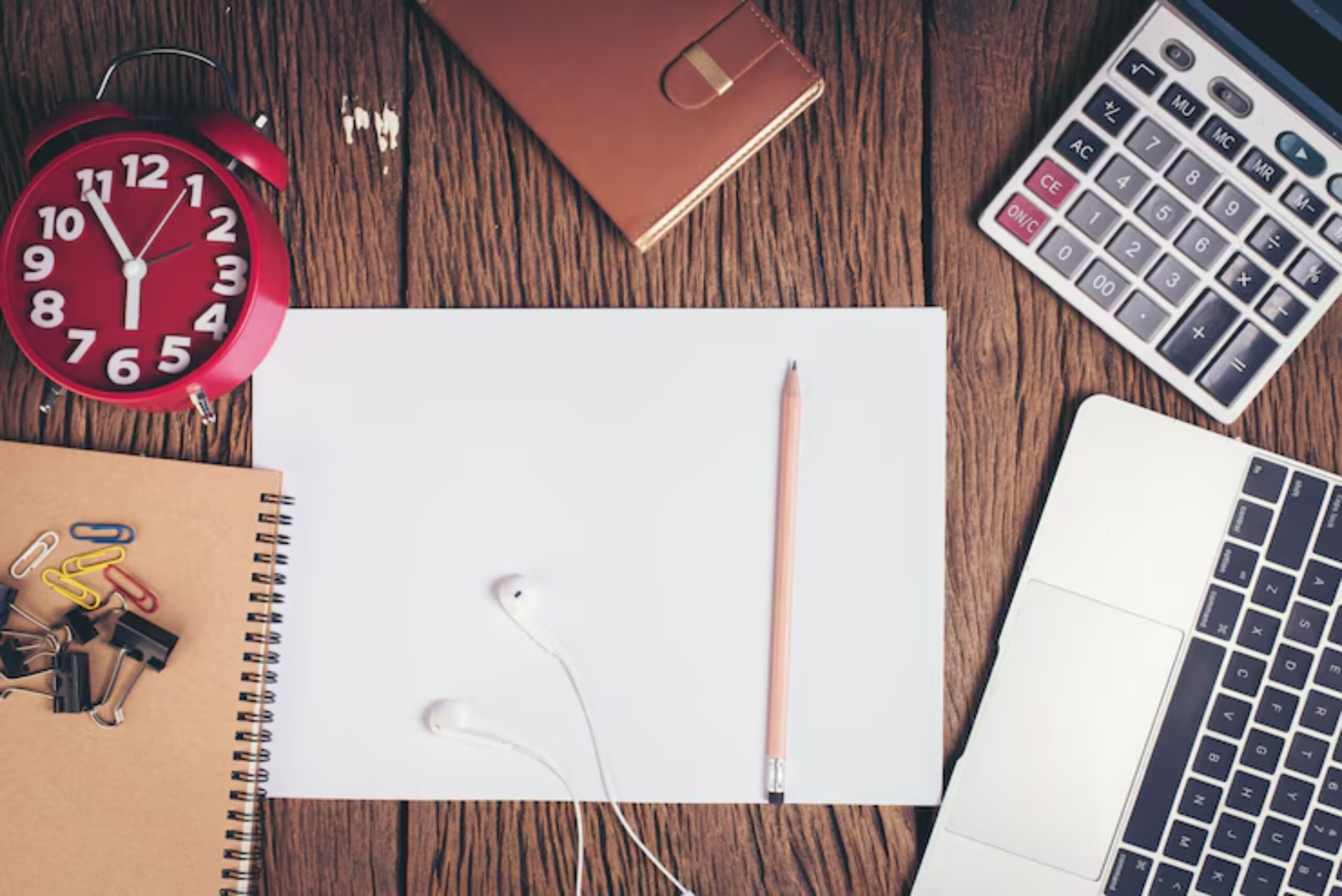 A workspace with a red alarm clock, notebook, pencil, earbuds, calculator, and blank paper on a wooden desk.
