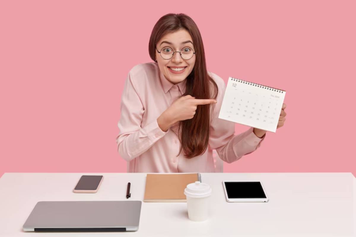 A person in a pink shirt points at a calendar on a desk with a laptop, notebook, pen, smartphone, and coffee cup in a pink background.