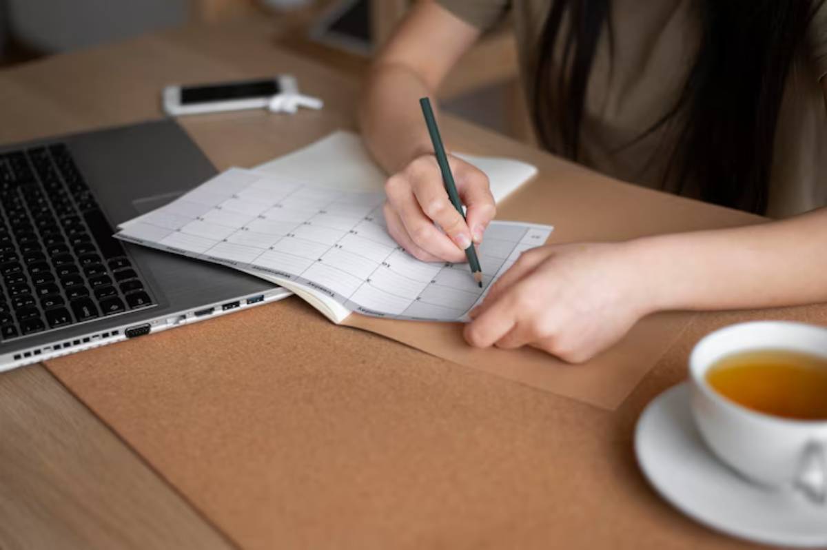 A person writing on a calendar while seated at a desk with a laptop, a cup of tea, and a smartphone nearby.