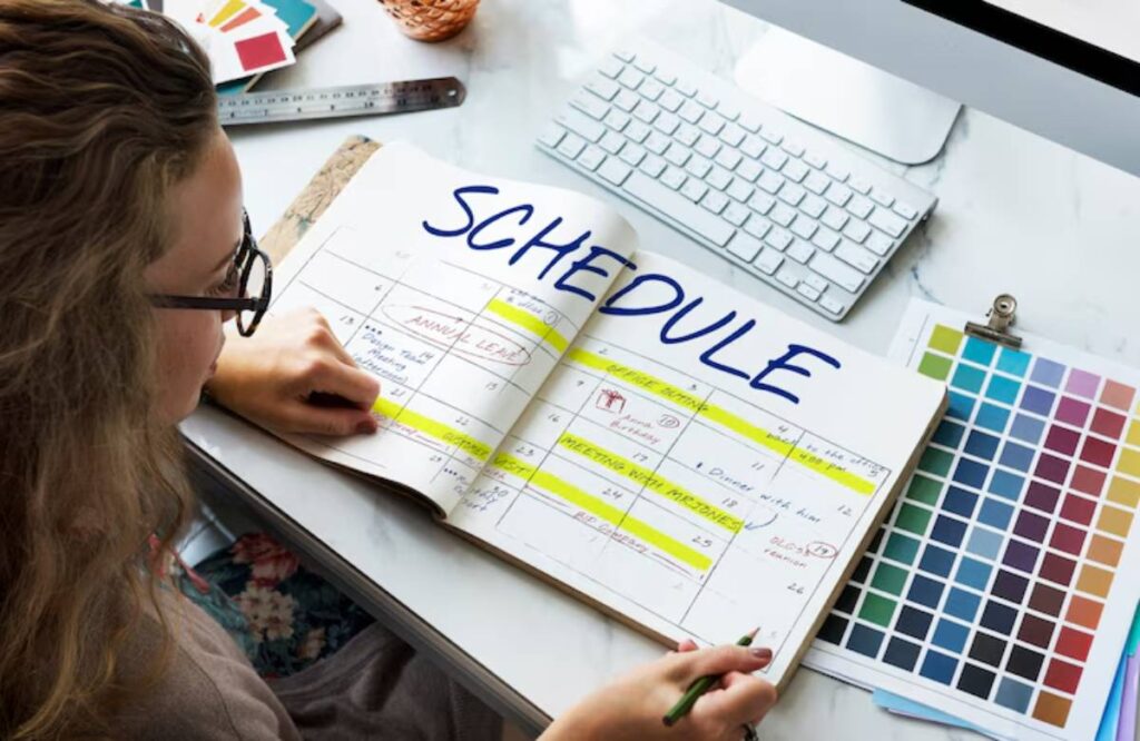 A hand writes in a planner labeled SCHEDULE, with highlighted dates and a computer alongside various color swatches in the background.
