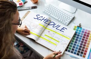 A hand writes in a planner labeled SCHEDULE, with highlighted dates and a computer alongside various color swatches in the background.