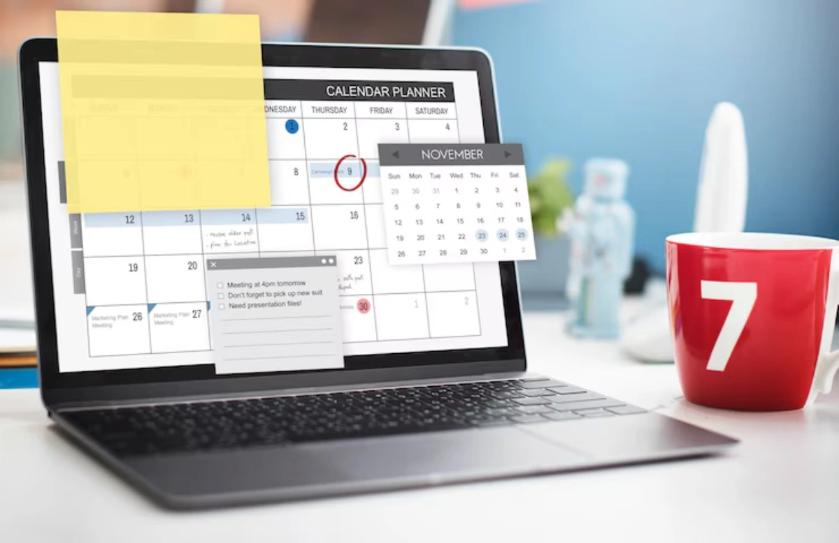 A laptop displaying a calendar planner with sticky notes and reminders for meetings, alongside a bright red mug.