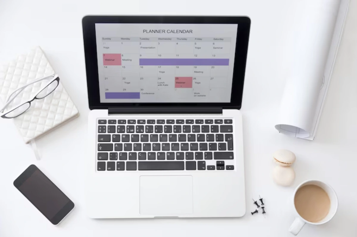 A laptop displaying a planner calendar, alongside glasses, a notebook, a phone, coffee, and macarons on a white desk.