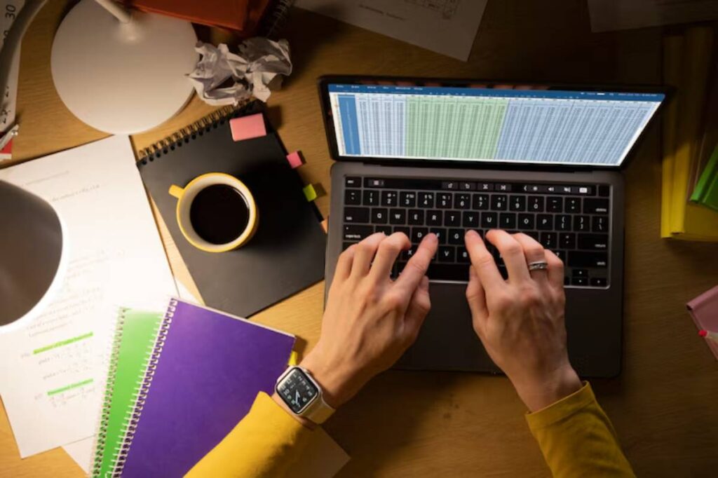 A person types on a laptop displaying a spreadsheet, surrounded by notebooks, a coffee mug, and crumpled paper, under warm light.