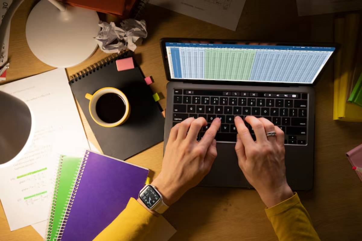 A person types on a laptop displaying a spreadsheet, surrounded by notebooks, a coffee mug, and crumpled paper, under warm light.