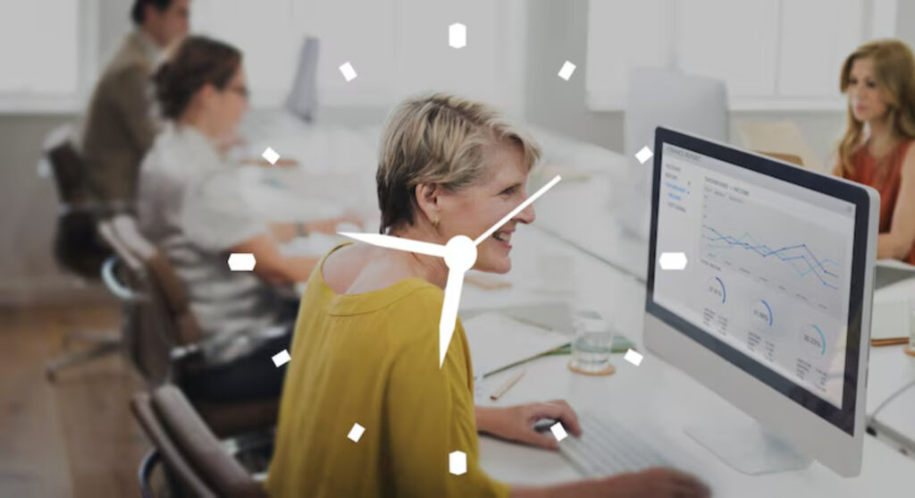 A woman in a yellow shirt works on a computer displaying financial data, surrounded by colleagues in a bright office setting.
