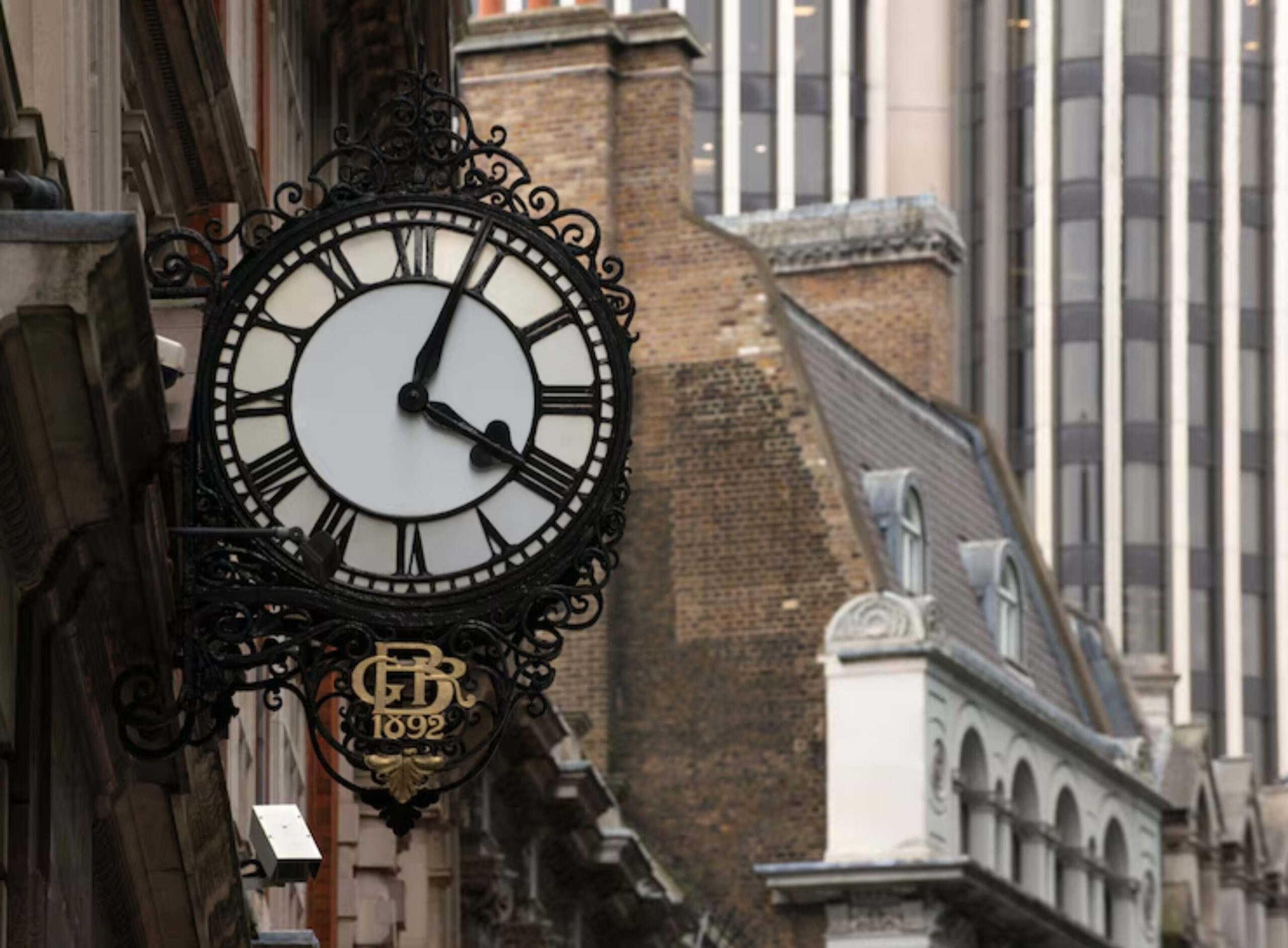 A vintage clock with Roman numerals hangs from a building, displaying ornate metalwork and the inscription "GR 1892."