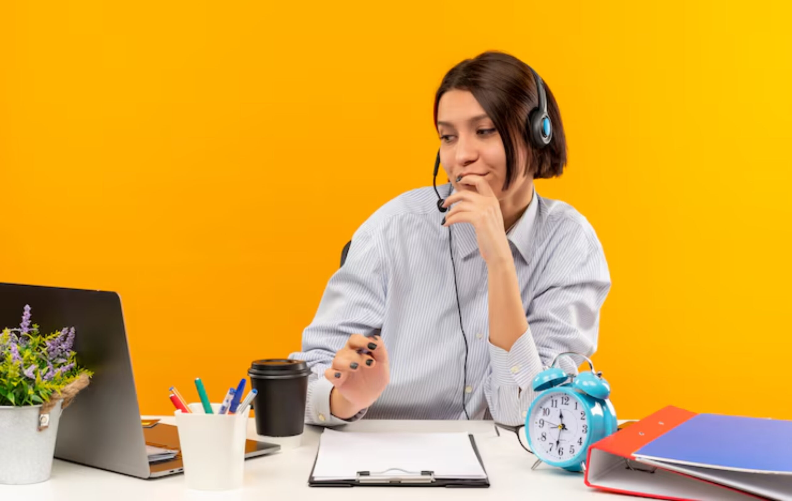 A person in a striped shirt wearing headphones sits at a desk with a laptop, notepad, coffee, and colorful stationery against a bright orange background.