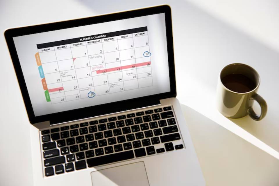 A laptop displays a monthly calendar with scheduled events, alongside a steaming cup of coffee on a white table.