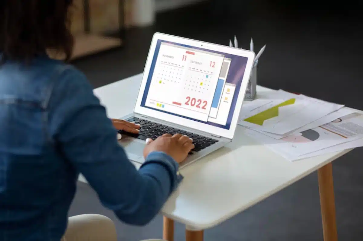 A person types on a laptop displaying a calendar for November and December 2022, surrounded by papers and stationery on a table.