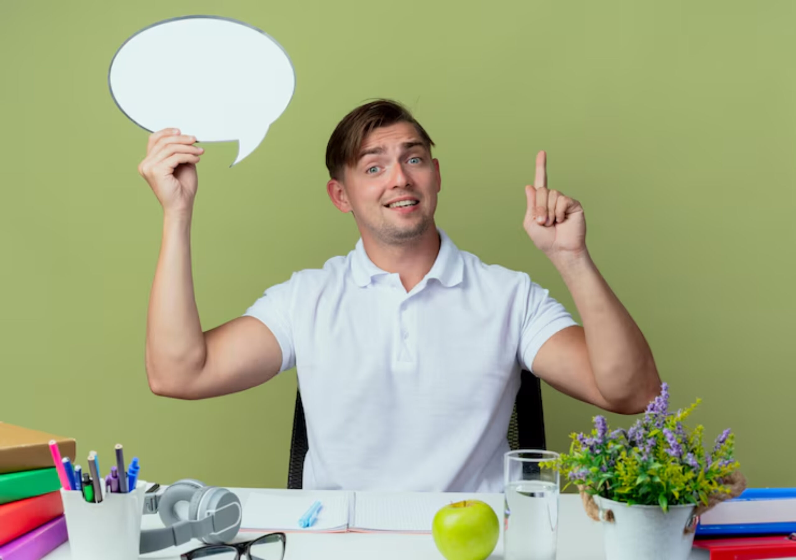 A person holding a speech bubble above their head, sitting at a desk with stationery, an apple, and a potted plant, against a green backdrop.