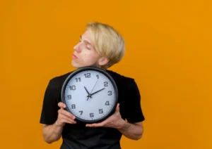 A person with blond hair holds a large wall clock in front of them, set against a bright orange background.