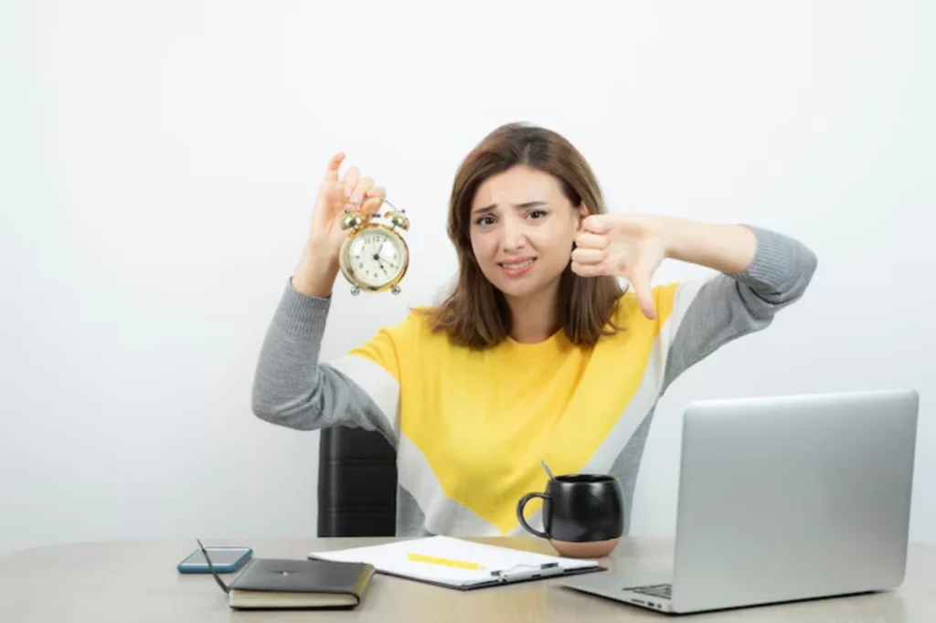 A woman in a yellow and gray sweater holds an alarm clock in one hand and gives a thumbs down with the other, seated at a desk.