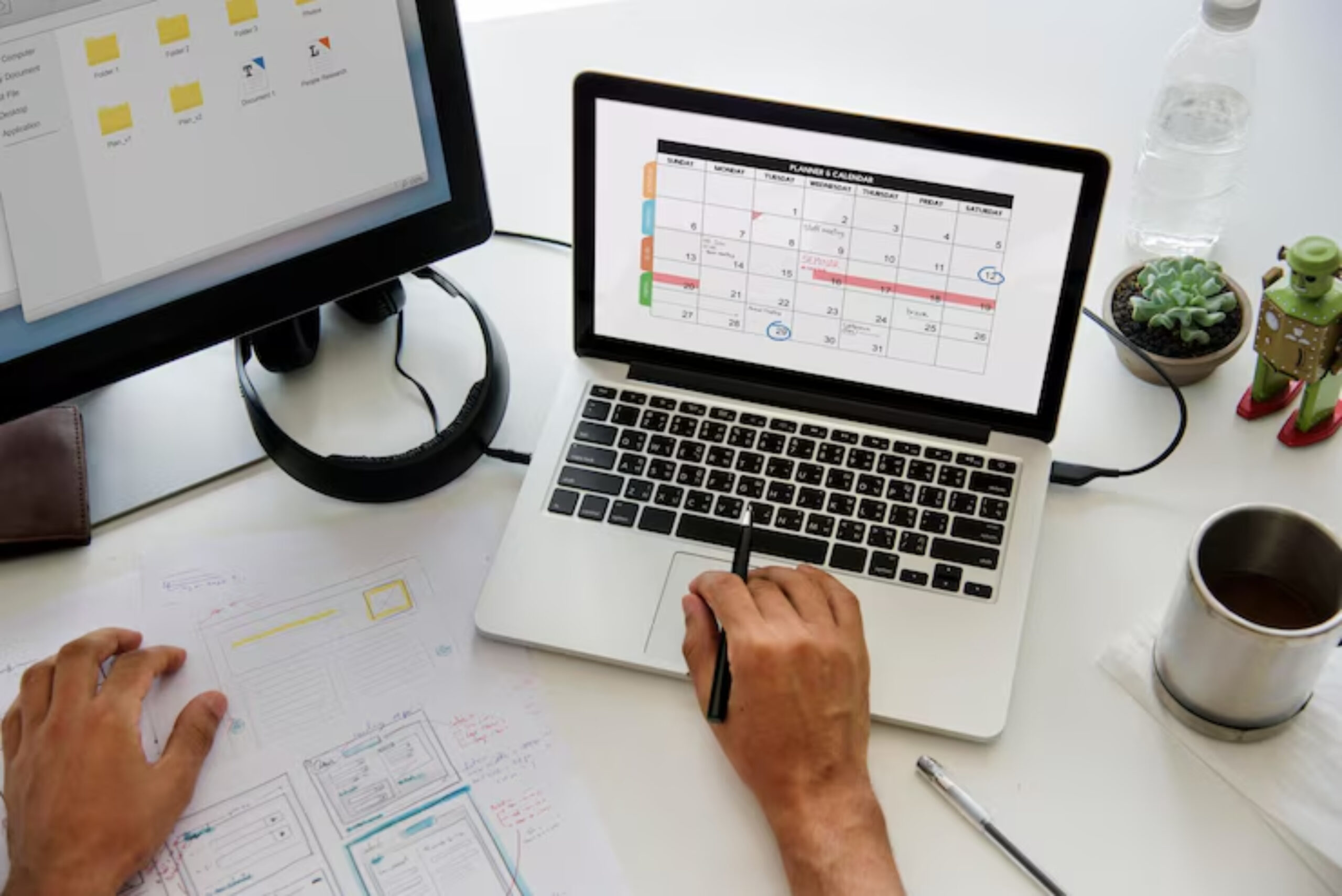 A workspace showing a laptop with a calendar, a hand with a pen, sketches, a mug, and a water bottle on a desk.