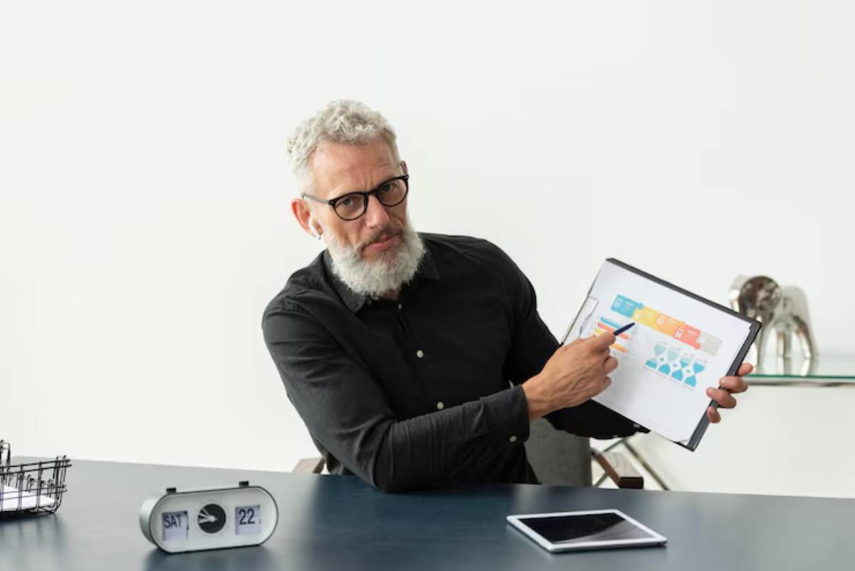 A person in a black shirt points to a chart on a clipboard during a presentation, with a clock and tablet on the desk.