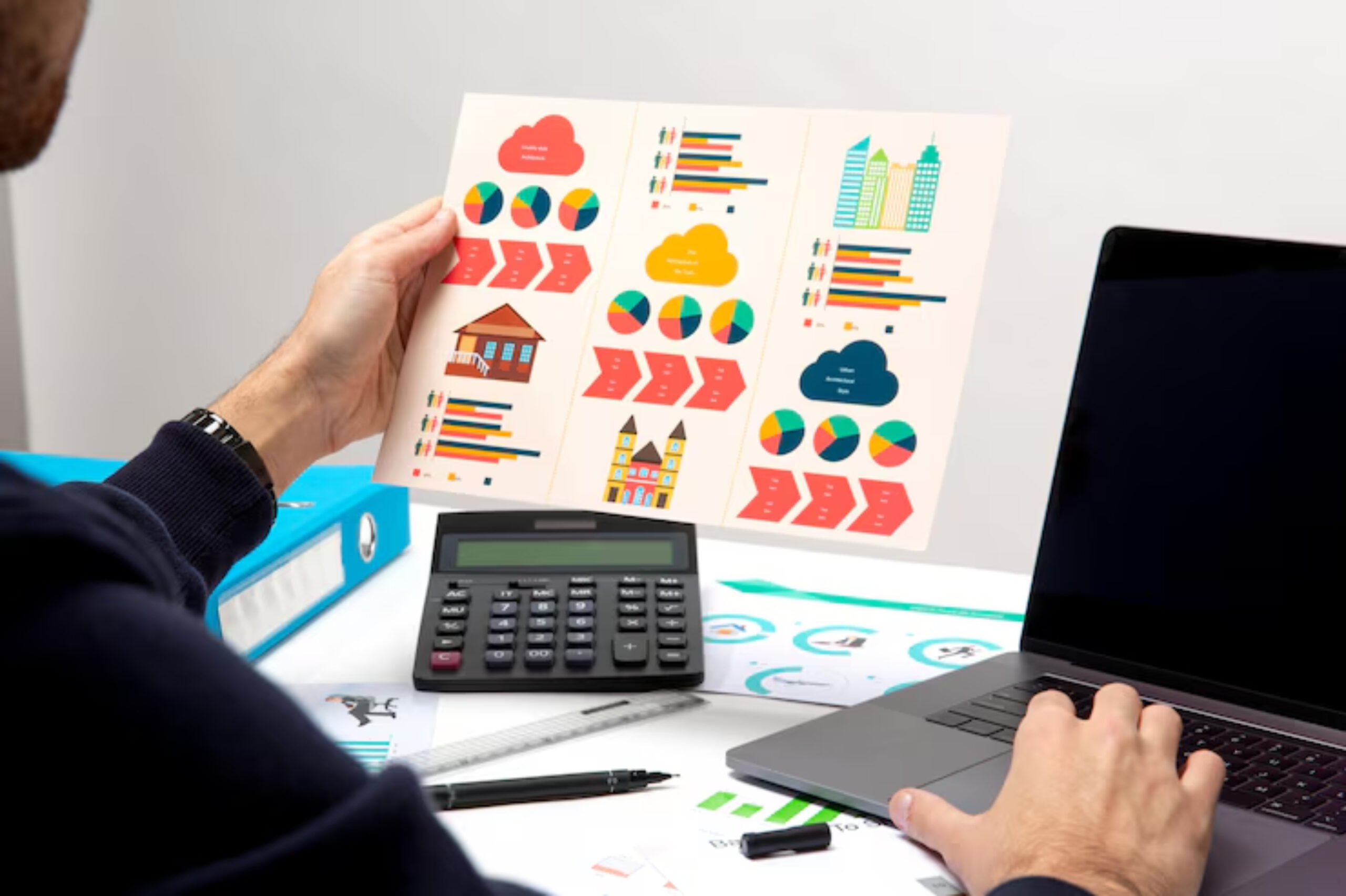 A person reviewing colorful charts and graphs, holding a report while seated at a desk with a laptop and calculator.