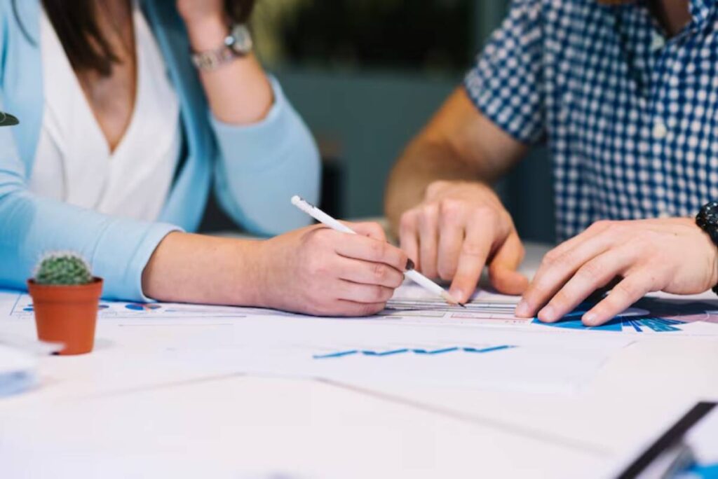 Two people collaborate on a project at a table, examining charts and documents, with one writing on paper while the other gestures.