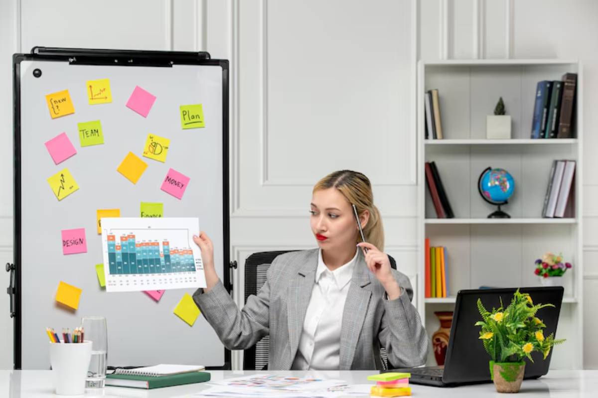 A professional woman in a gray blazer holds a chart while sitting at a desk with a laptop, plants, and sticky notes behind her.