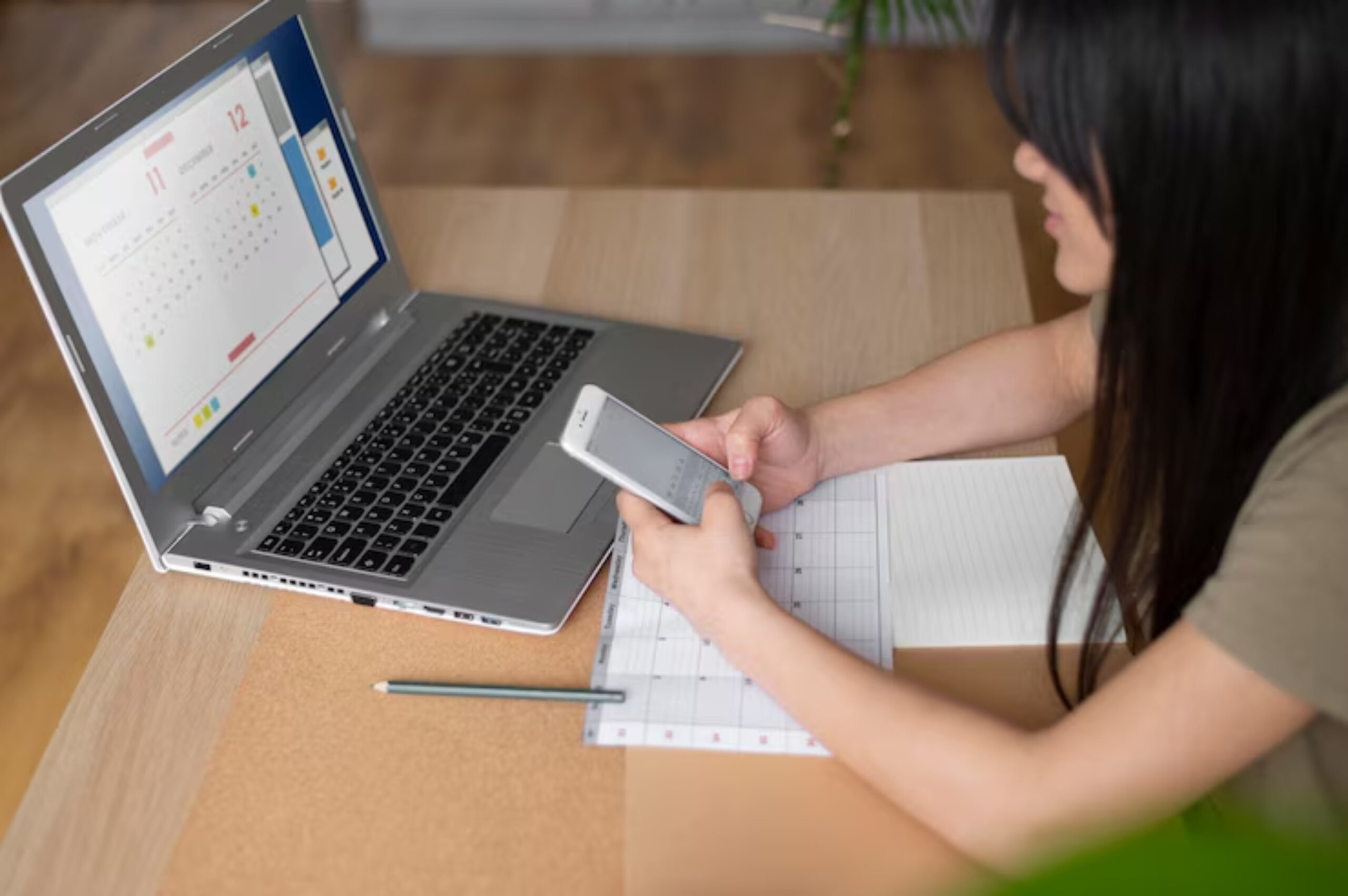 A person holds a smartphone while sitting at a desk with a laptop displaying a calendar and a notepad with a pencil nearby.