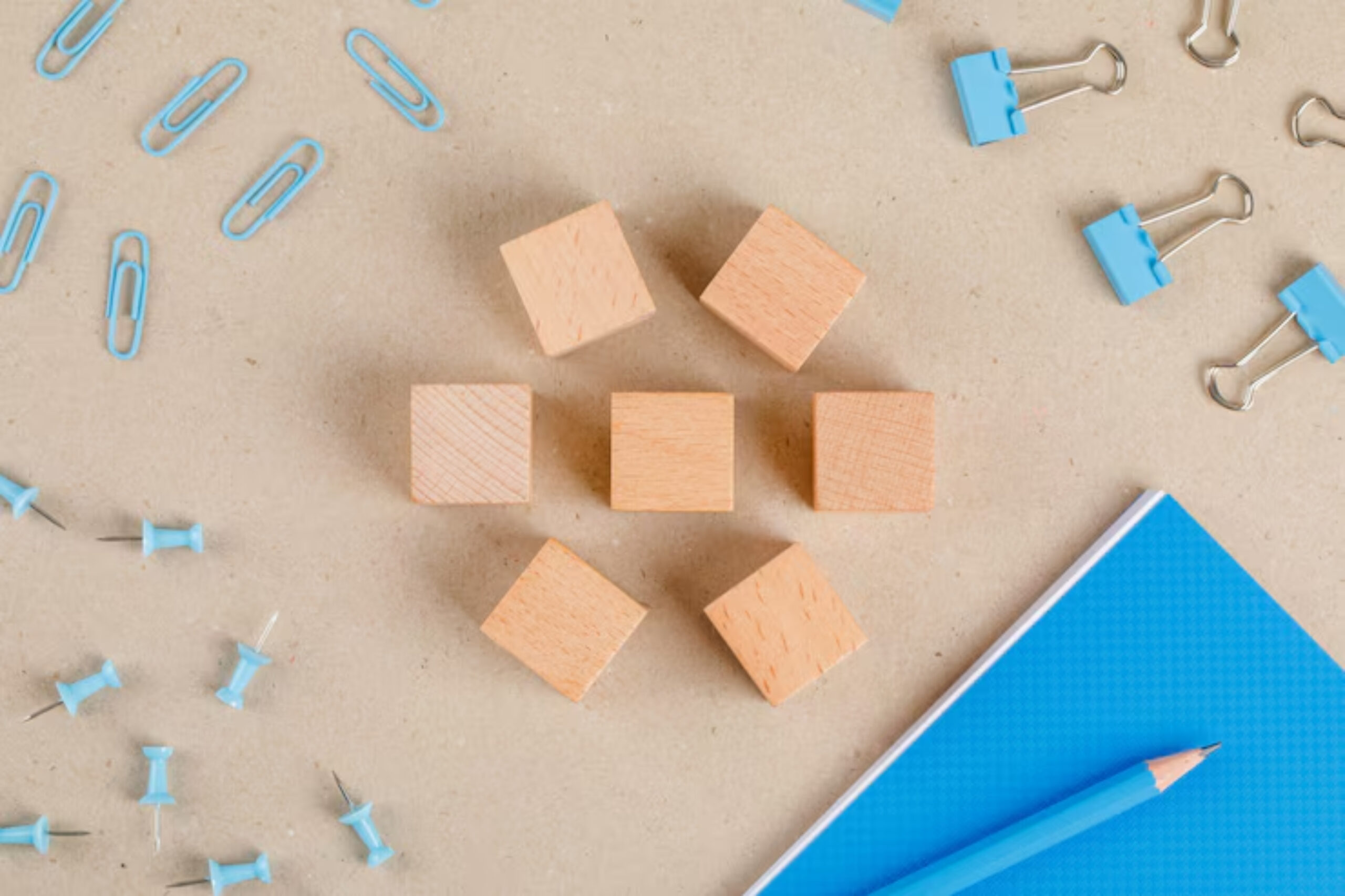 A flat lay of wooden cubes, blue paper clips, push pins, a blue notebook, and a pencil on a textured brown surface.