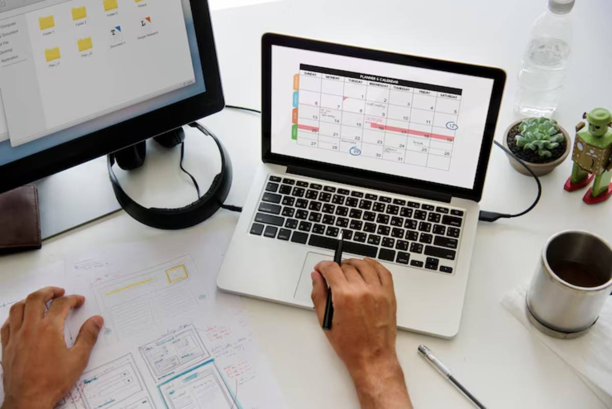 A person uses a laptop with a calendar on the screen while taking notes on paper, surrounded by a water bottle, coffee, and a plant.