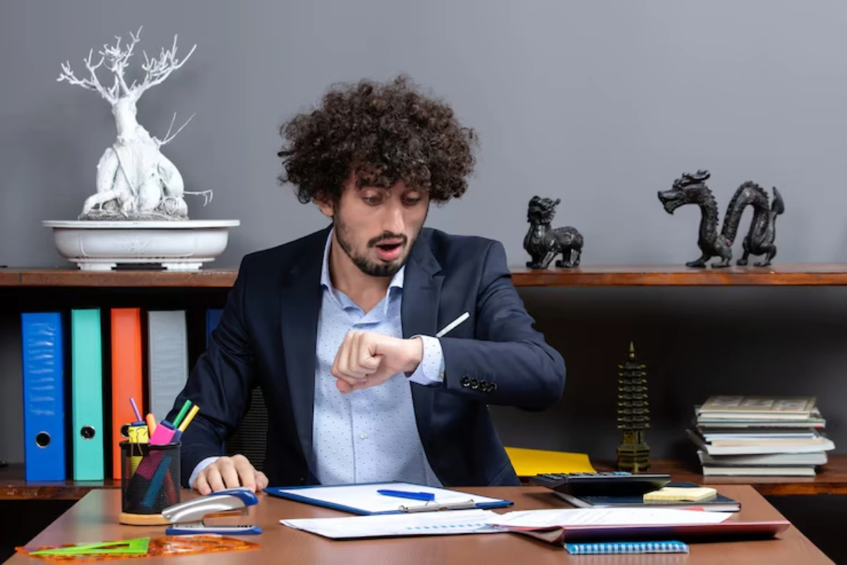 A person in a business suit checks their watch while sitting at a desk surrounded by stationery, books, and decorative items.