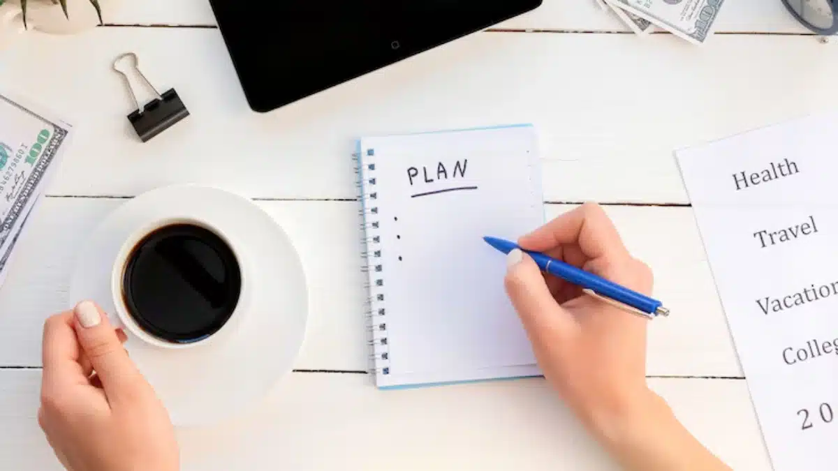 A hand writing PLAN in a notebook beside a cup of coffee, money, a tablet, and a paper with various goals listed.