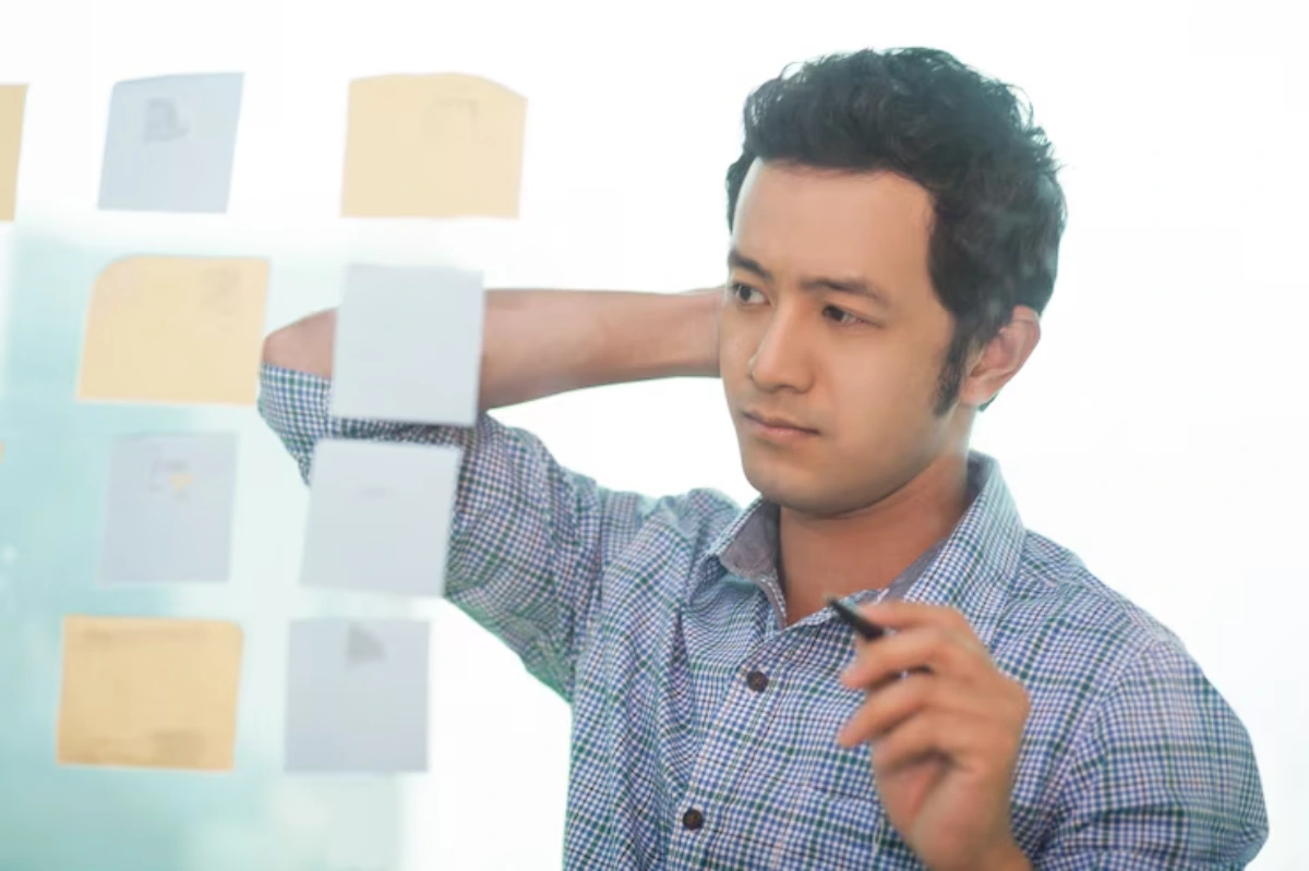 A person in a checkered shirt stands by a glass wall covered with colorful sticky notes, holding a pen and deep in thought.