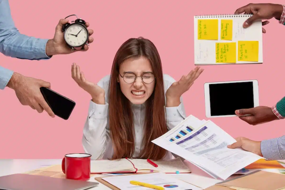 A woman appears overwhelmed surrounded by a clock, smartphone, tablet, and printed documents on a cluttered desk against a pink background.