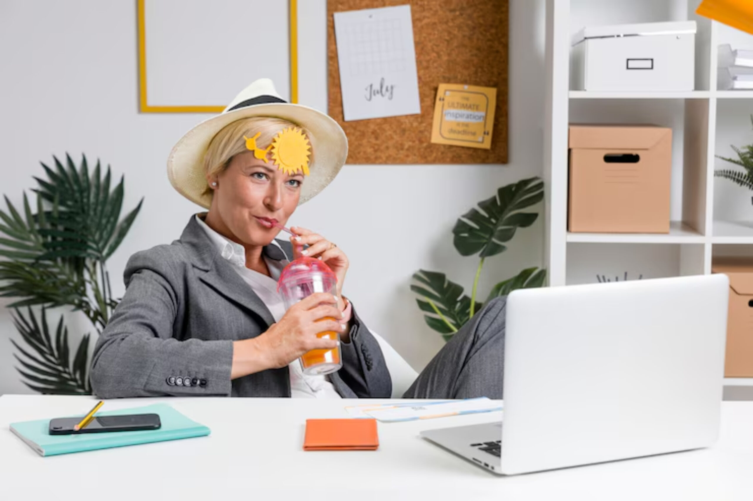 A person in a stylish gray suit and wide-brimmed hat relaxes in a modern office, sipping from a colorful drink.