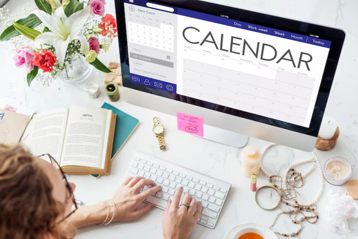 Overhead view of a desk with a computer displaying a calendar, an open book, flowers, and various accessories. A woman types on a keyboard.