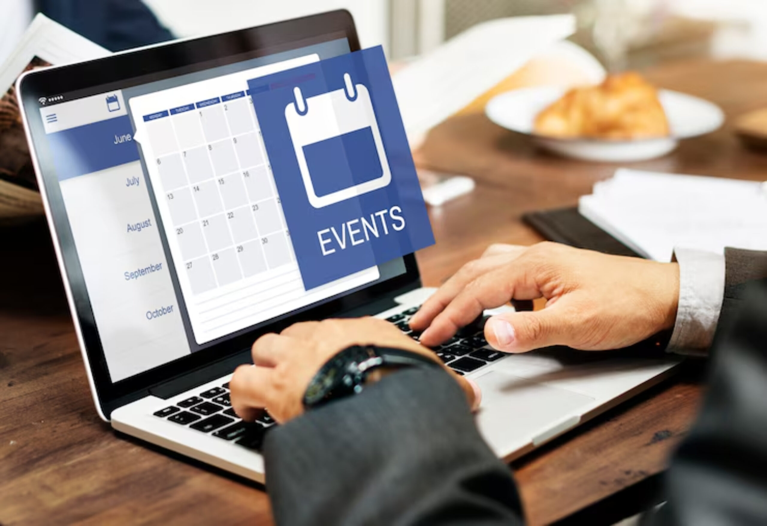 A person using a laptop to manage events on a digital calendar, with a plate of food in the background.