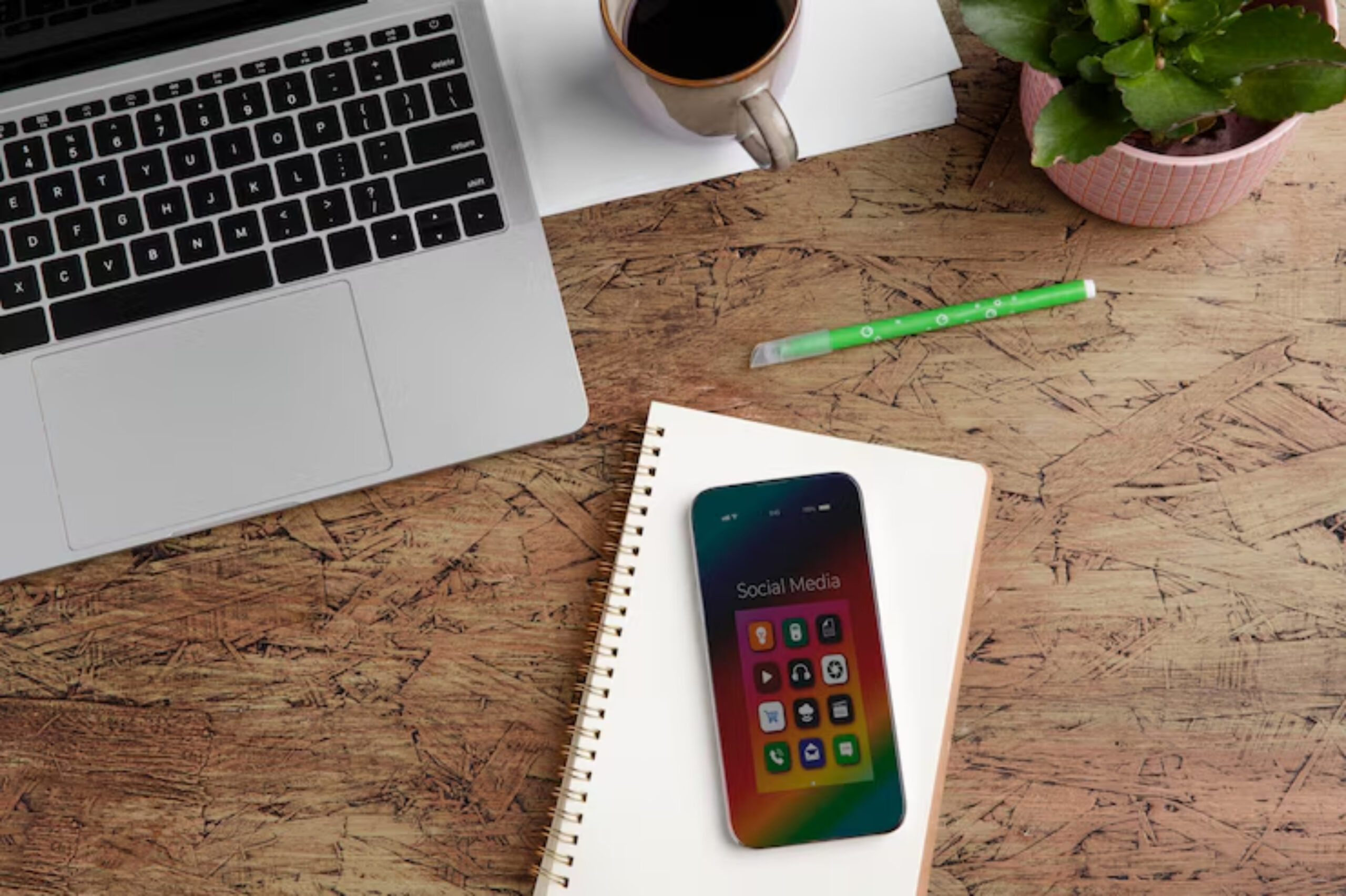 A smartphone displaying social media icons rests on a notebook, alongside a laptop, coffee, a notepad, and a green pen on a wooden desk.