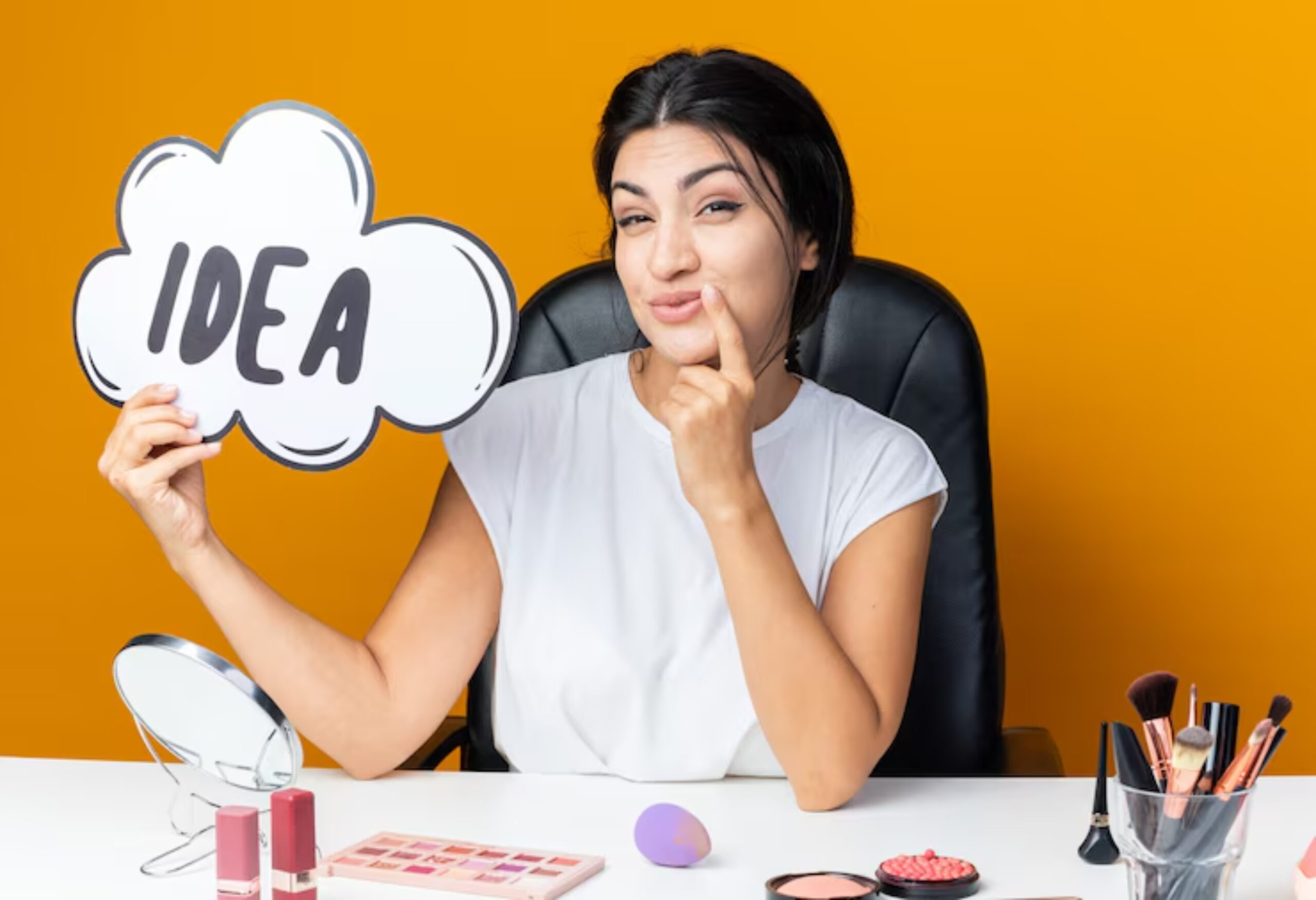 A person sits at a vanity desk holding a speech bubble sign that says IDEA, surrounded by makeup products on a bright yellow background.