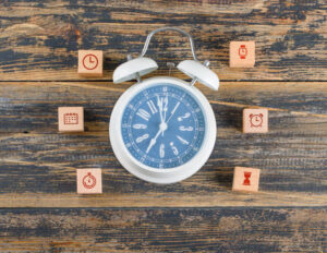 A white alarm clock shows 10:10, surrounded by six wooden blocks with time-related icons, on a rustic wooden surface. The scene conveys themes of time management.