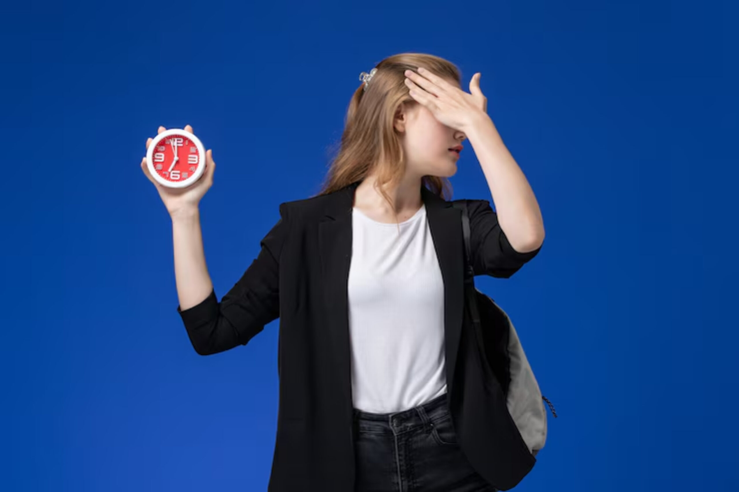 A person in a black blazer and white shirt stands against a blue background, holding a red clock in one hand.