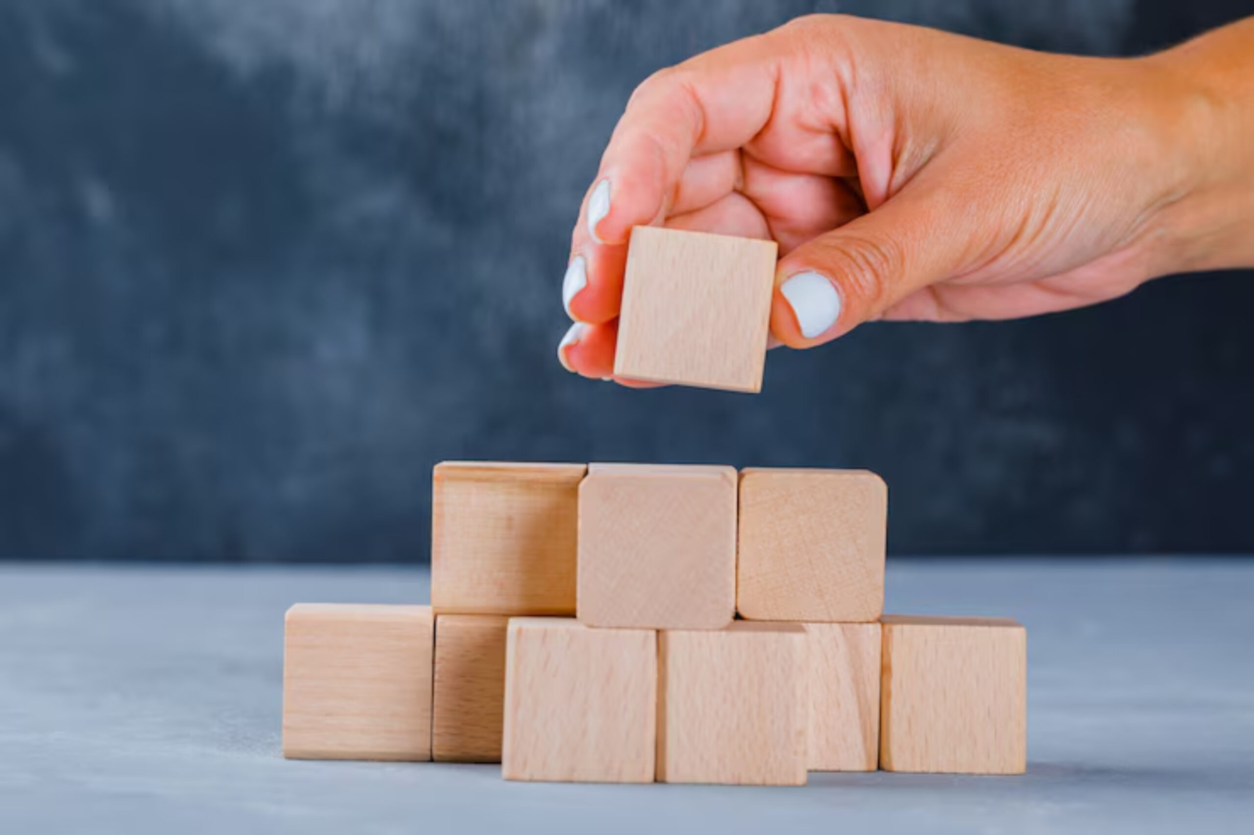 A hand is stacking a wooden block on top of a pyramid of similar blocks against a blurred blue backdrop.