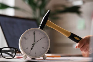 A hand poised to hit a white alarm clock with a yellow-handled hammer, set on a desk beside glasses and a laptop.