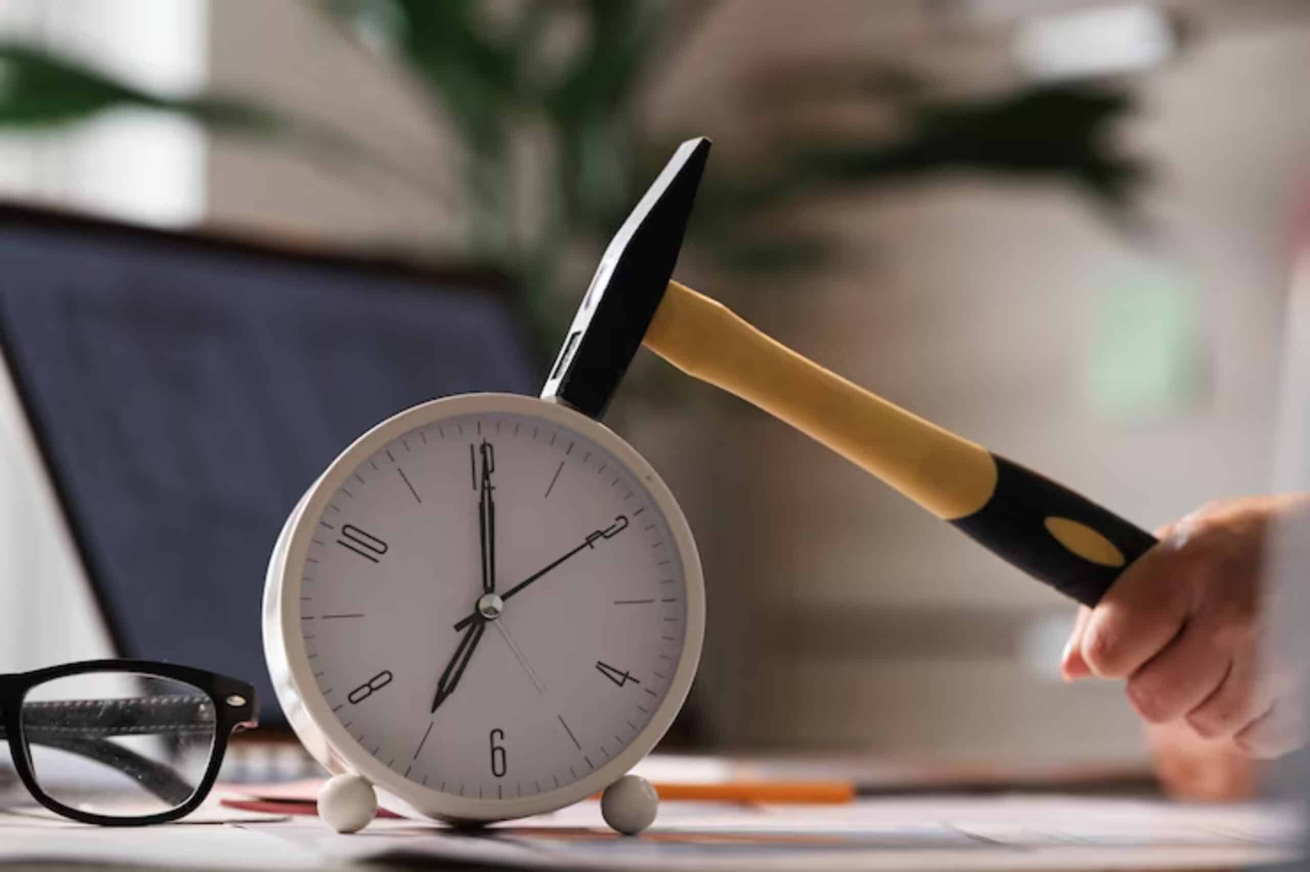 A hand poised to hit a white alarm clock with a yellow-handled hammer, set on a desk beside glasses and a laptop.