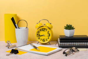 A workspace featuring a yellow alarm clock, notebooks, pens, a planter, and stationery on a pastel surface against a yellow background.