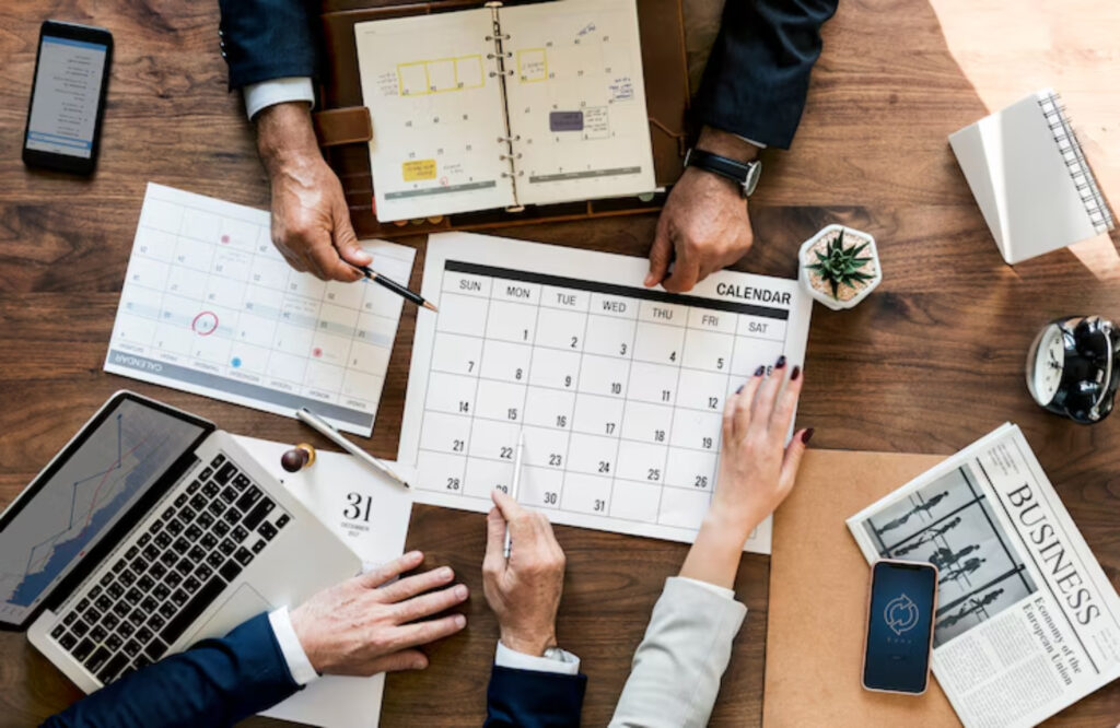 Hands of business professionals gather around a calendar and laptop, planning schedules amid paperwork and a potted plant on a wooden table.