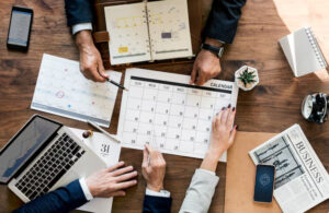 Hands of business professionals gather around a calendar and laptop, planning schedules amid paperwork and a potted plant on a wooden table.