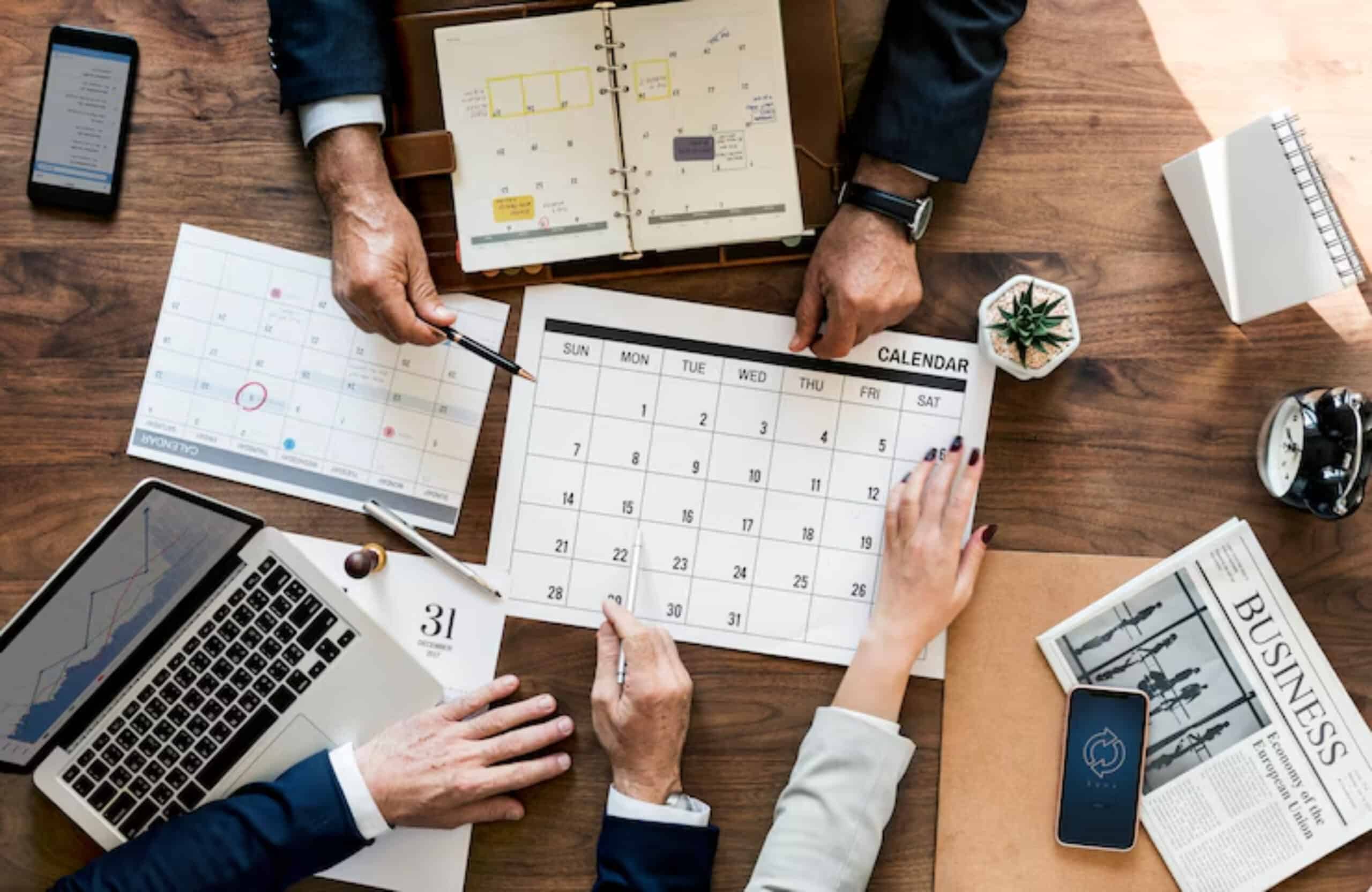 Hands of business professionals gather around a calendar and laptop, planning schedules amid paperwork and a potted plant on a wooden table.