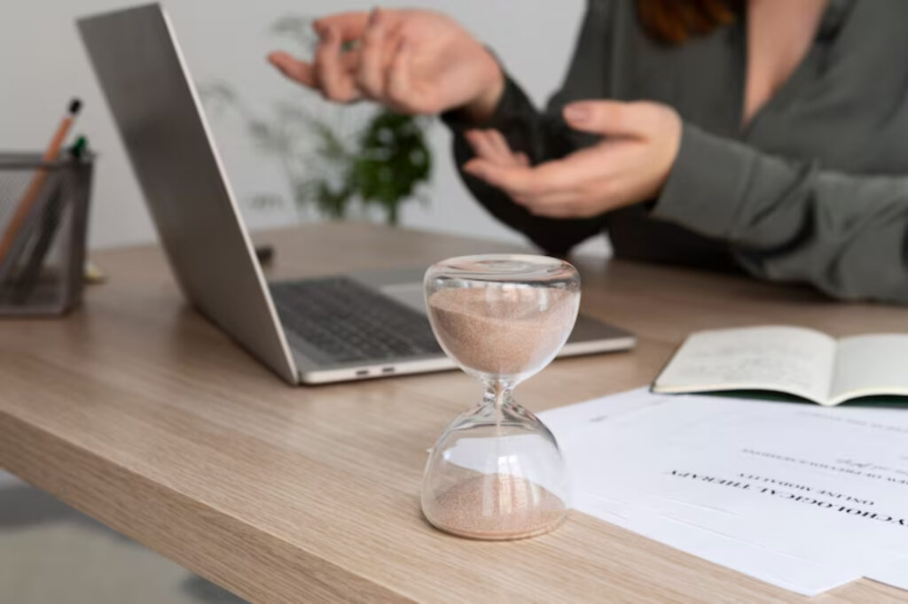 A person gestures expressively while discussing something at a desk with a laptop and an hourglass, symbolizing time and urgency.
